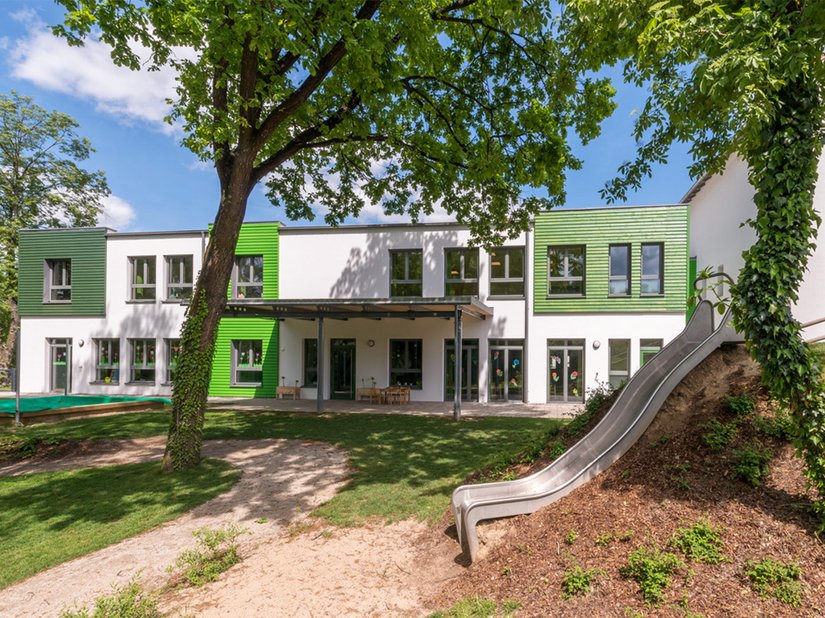 Grüne Holzverkleidungen lockern die Gestaltung der Außenfassade optisch auf. Die komplementäre Farbgebung grenzt die Kindertageseinrichtung von der benachbarten Grundschule mit roten Elementen ab.