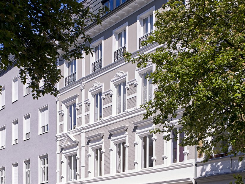 Die Stilfassade erstrahlt in einem warmen, den Farbton Taupe ähnlichen Grauton. Die stark gegliederten und verzierten Fenstergewände sind in Weiß gefasst.