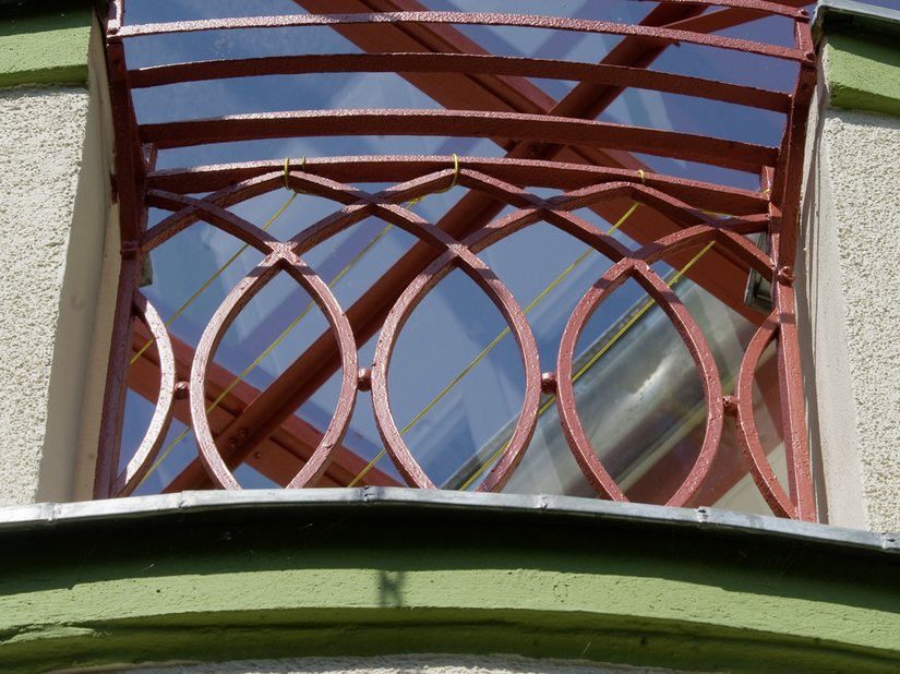 <p>Ein rotes Metallgeländer rundet mit der Tragekonstruktion des Glasdaches über dem halbrunden Vorbau des Vordachs ab.</p>