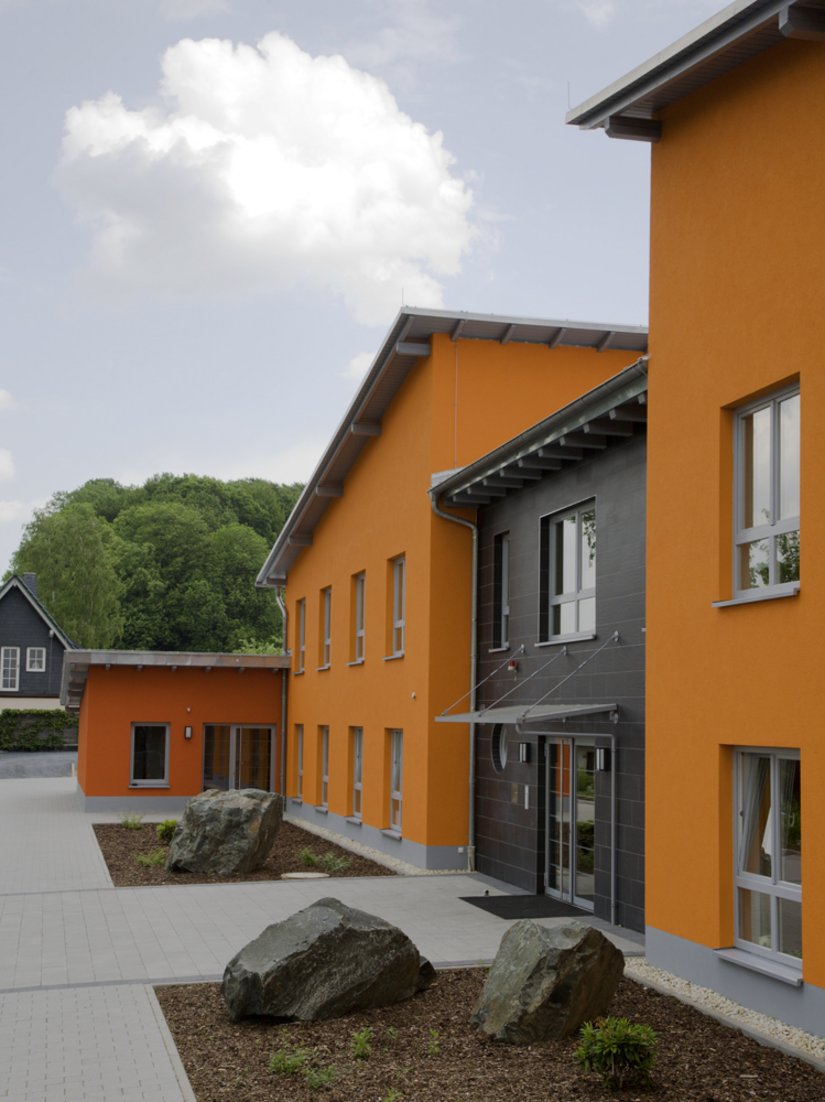 <p>Die eingeschossigen Eckgebäude mit dem dunklen orangeroten Farbton wirken wie eine Einfassung und lassen die einzelnen Baukörper als ein geschlossenes Ganzes erscheinen.</p>