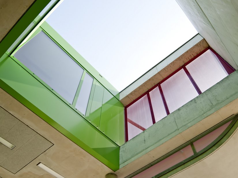 <p>Die Lichthöfe im Inneren sind als grün lackierte metallene „Futterale“ ausgebildet. Die Betonoberflächen der Einschnitte der Außenräume sind dagegen betont rau strukturiert und partiell durch den vierlagigen Auftrag einer roten Lasur akzentuiert.</p>